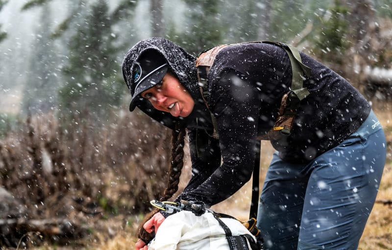 Backpacker adjusting gear in a snowstorm, wearing a black hoodie and carrying a white pack.