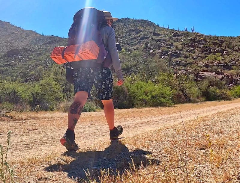  Person hiking a sunny desert trail with a large backpack, wearing Wild Rye shorts and an orange foam pad strapped to the pack.