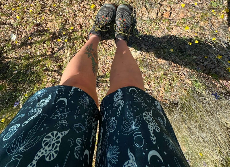  Top-down view of someone wearing Wild Rye printed shorts and trail runners next to two cameras on rocky desert ground.