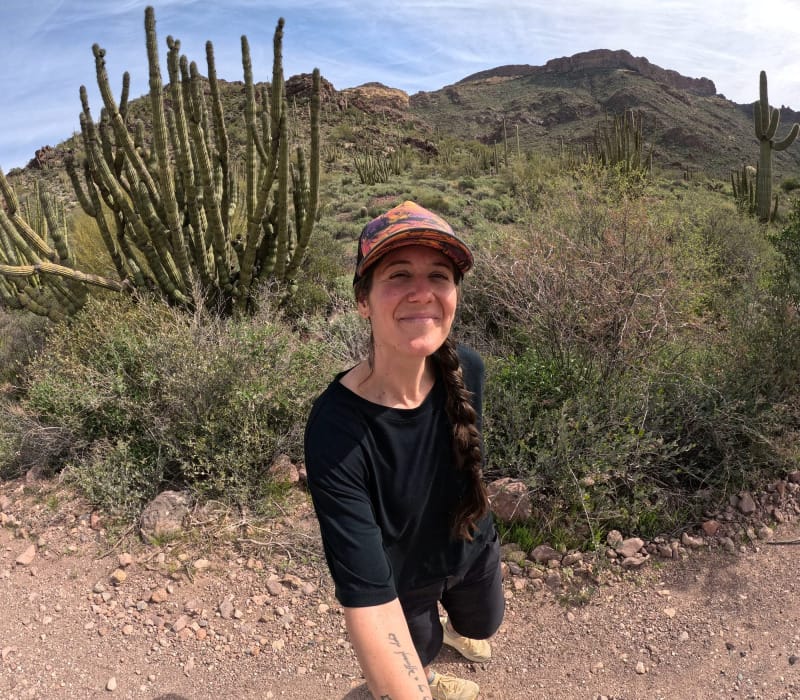  Smiling woman in black Wild Rye top and colorful cap hiking among cacti in the Sonoran Desert.