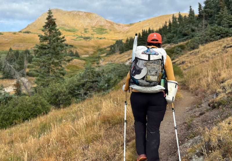 Hiker with trekking poles carrying the Hyperlite Windrider 70 Backpack on an alpine trail surrounded by golden meadows.