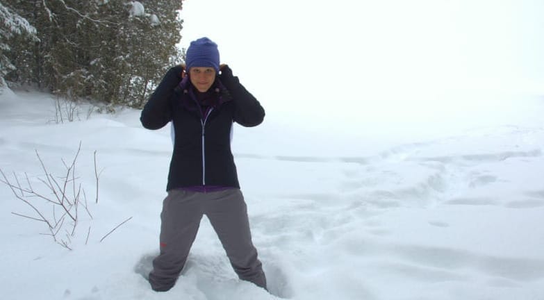 Hiker in purple hat and black jacket standing in deep snow with evergreen trees.