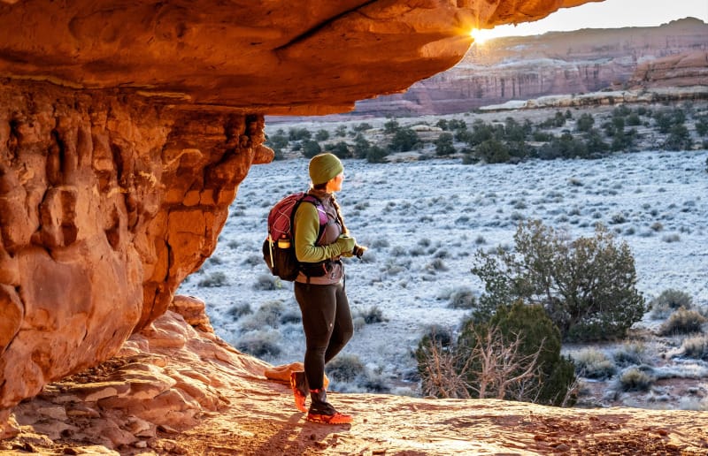 Hiker with backpack standing under canyon overhang at sunrise with frosty desert landscape.