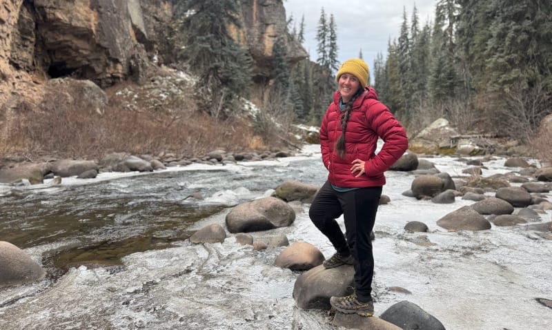 Hiker standing on icy river rocks wearing a red insulated jacket and yellow beanie.
