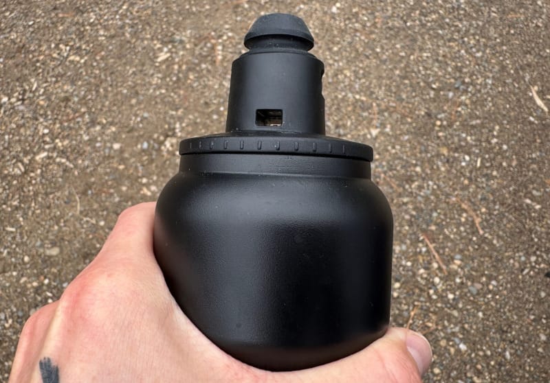 Hand holding the Tiibo Vacuum Insulated Bottle with a close-up view of the black nozzle-style lid against a dirt trail background.
