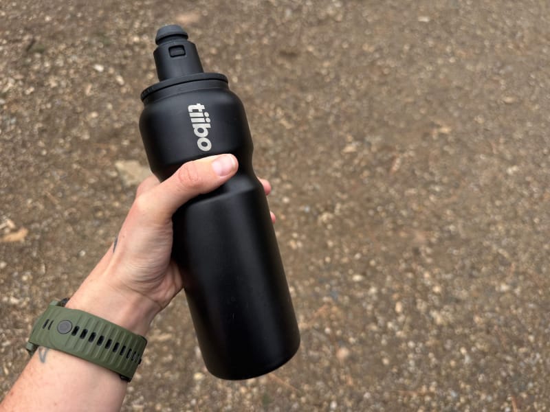 Hiker holding a black Tiibo Vacuum Insulated Bottle with logo visible while standing on a dirt trail.
