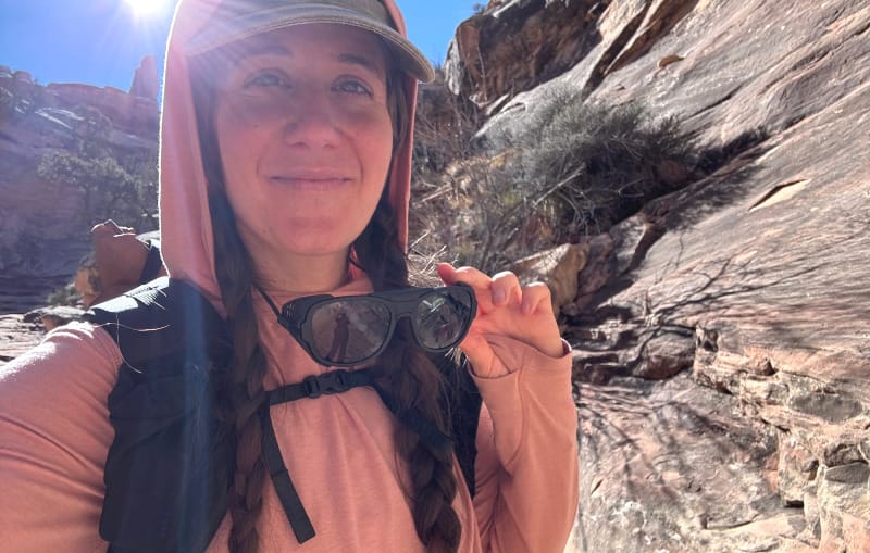Hiker in a canyon holding sunglasses, wearing layered cold-weather hiking clothes with sun overhead.