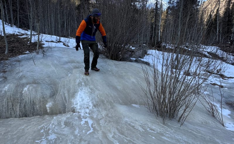 Hiker in orange jacket walking across icy trail surrounded by snow and trees.