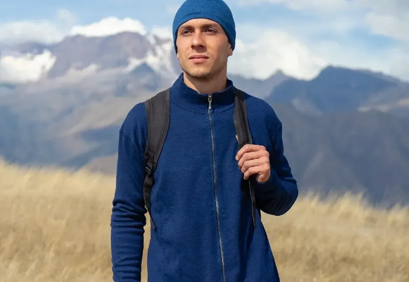 Hiker wearing a blue Arms of Andes alpaca wool zip-up and beanie standing in front of mountains in the Peruvian Andes.