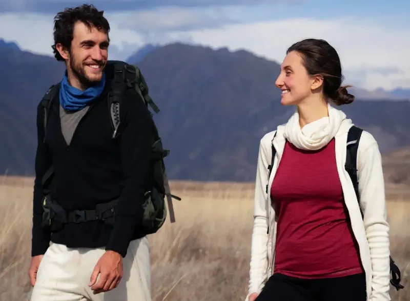 Two hikers wearing Arms of Andes alpaca wool clothing walking through a high-altitude grassland with mountain peaks in the background.