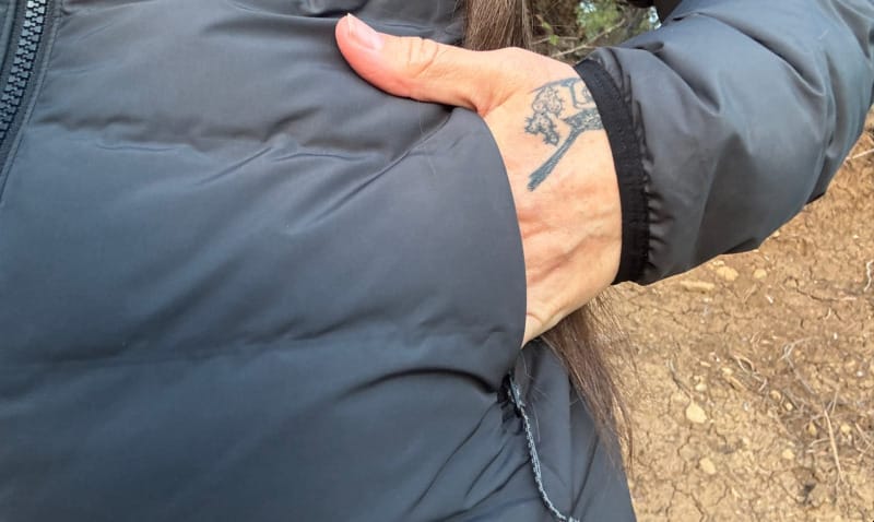 Close-up of a person’s hand inside the exterior pocket of the black KUIU Elements Hooded Jacket, showing durable zipper and stitching.
