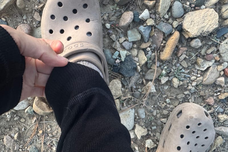 Close-up of a person adjusting the cuff of black PAKA Thermal Pants near hiking Crocs on rocky ground, showing snug ankle fit.