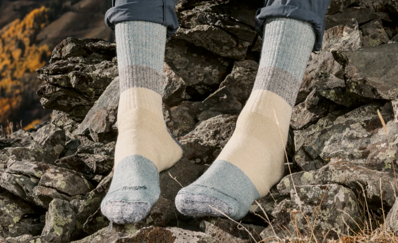 Hiker wearing neutral-toned Darn Tough socks while resting on rocks with fall colors in the background.