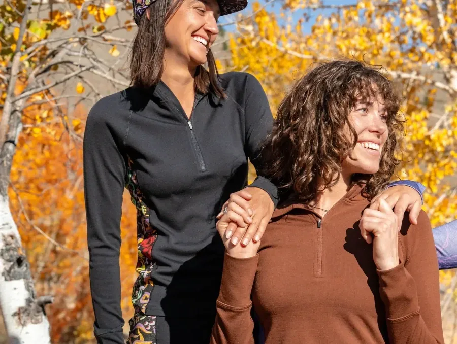 Two women wearing Wild Rye Bassett baselayers standing in bright autumn foliage, showing the half-zip top and patterned leggings.