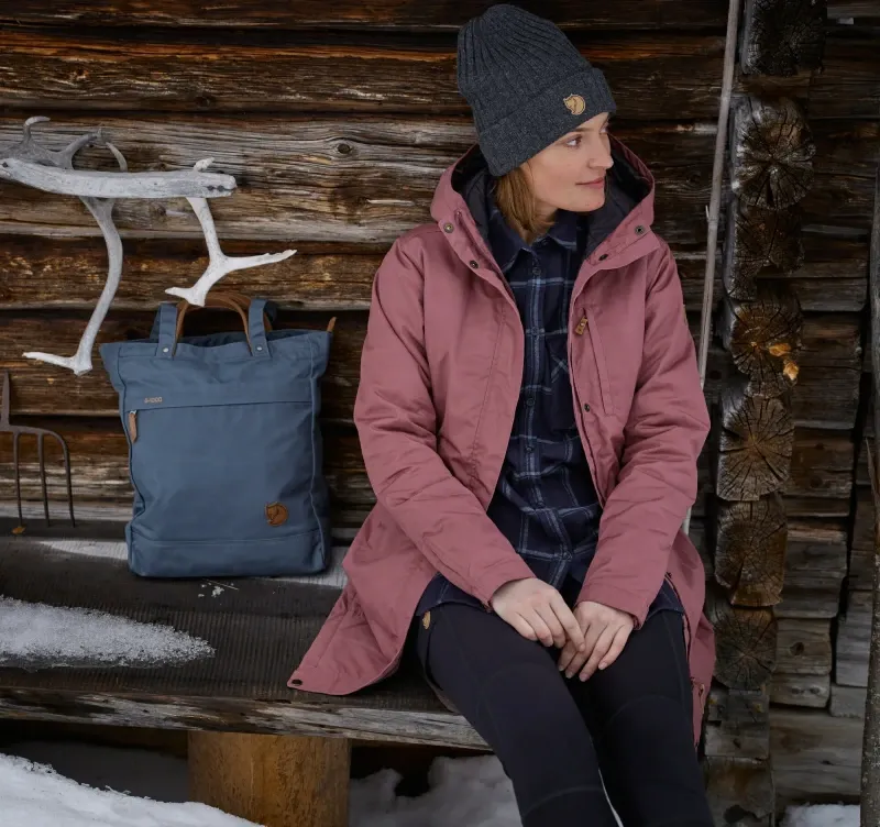 Woman sitting outside a rustic cabin wearing Fjallraven Abisko Trail Tights and winter layers in a snowy setting.