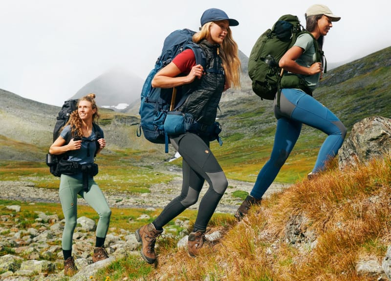 Three hikers wearing various Fjallraven Abisko trekking tights styles while climbing a grassy alpine slope with backpacks.
