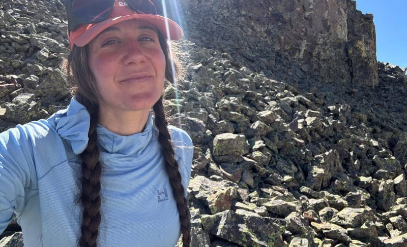 Hiker field testing a light blue sun hoodie on a rocky alpine trail in bright sunlight, assessing sun protection, breathability, and comfort during backpacking.