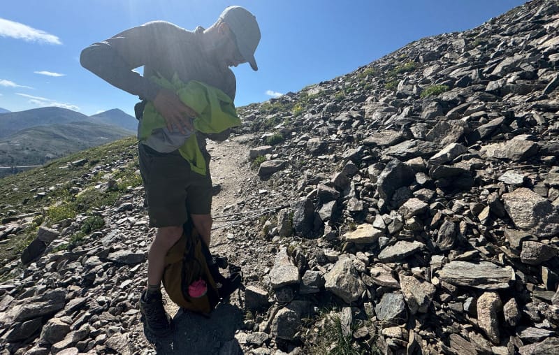 Hiker adjusting layers while wearing HIMALI clothing on a rocky alpine trail under clear blue skies