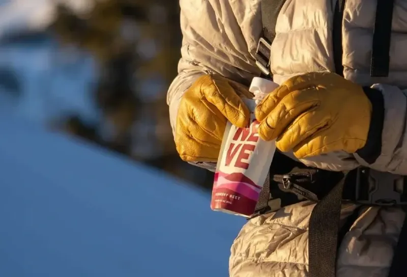 Hiker wearing a puffy jacket opening a Neve Foods smoothie pouch on a snowy trail during a winter hike.