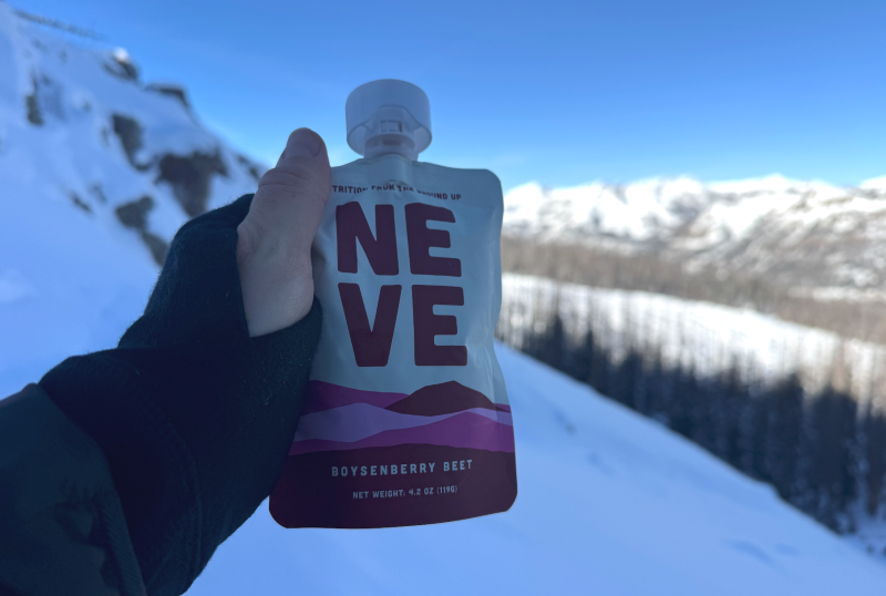 Hand holding a Neve Foods Boysenberry Beet smoothie pouch on a snowy mountain trail with alpine peaks in the background.