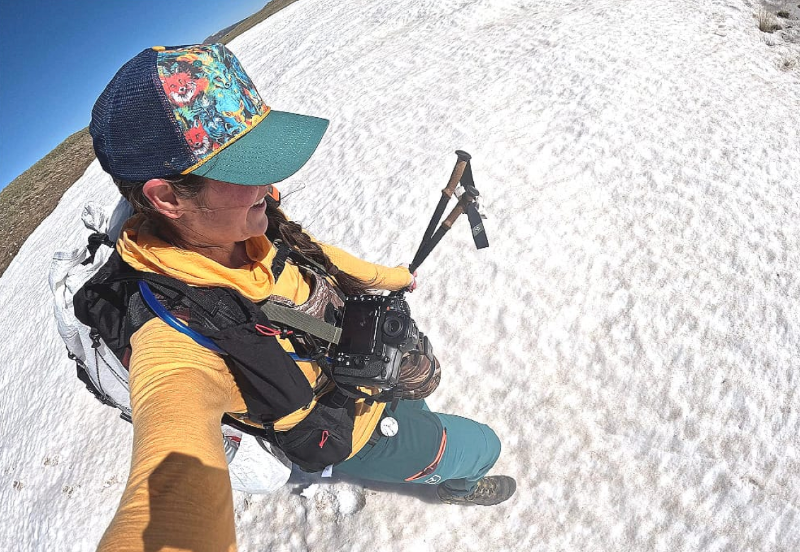 Person hiking across a snow-covered alpine slope wearing a white Outdoor Vitals CS40 Ultra Backpack, holding trekking poles and a camera, with spring snow covering the ground.