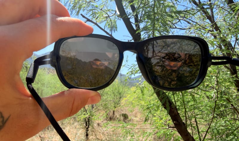 Ombraz sunglasses held up against a Sonoran Desert landscape showing the tinted lens view of mountains and desert vegetation.