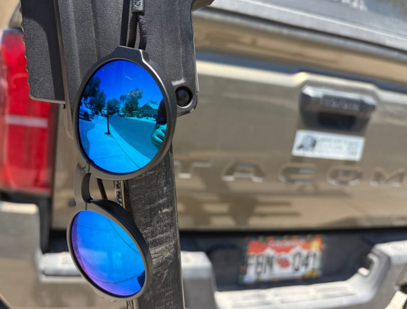 Ombraz Blue Mirror sunglasses hanging from outdoor gear in front of a dusty Toyota Tacoma.