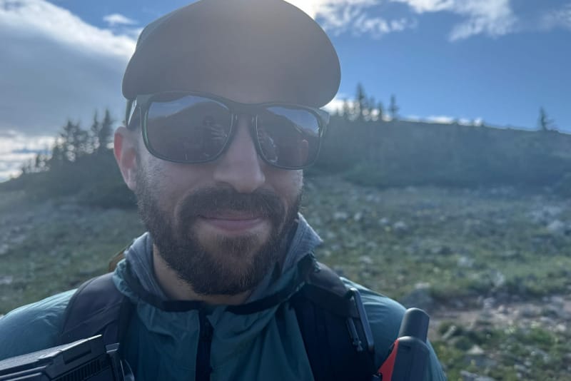 Hiker wearing Smith Guide's Choice sunglasses in an alpine environment with mountains, forest, and bright sunlight in the background.
