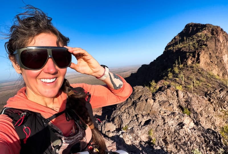 Hiker wearing Vuarnet sunglasses while standing on a rocky desert ridgeline with saguaro-covered mountains in the background.