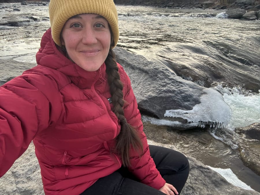 Hiker wearing a red down jacket and yellow knit hat beside an icy mountain stream in winter.
