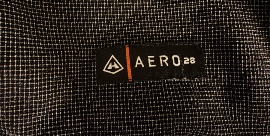 Detailed close-up of the Hyperlite Mountain Gear Aero 28 label stitched onto the black grid-pattern mesh exterior pocket.