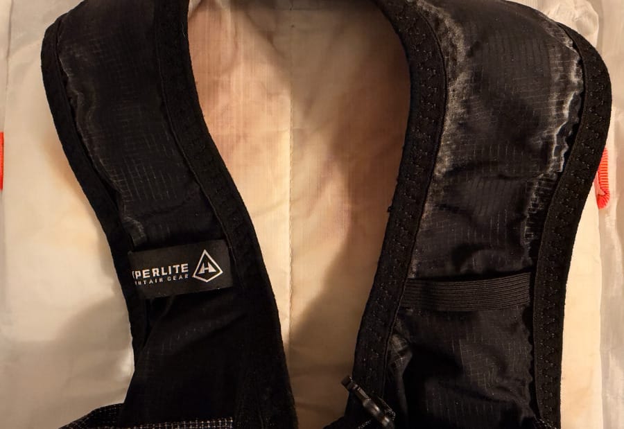 Close-up of the Hyperlite Mountain Gear Aero 28 running vest-style shoulder harness showing black fabric, padded edging, stitching, and the white backpack body.