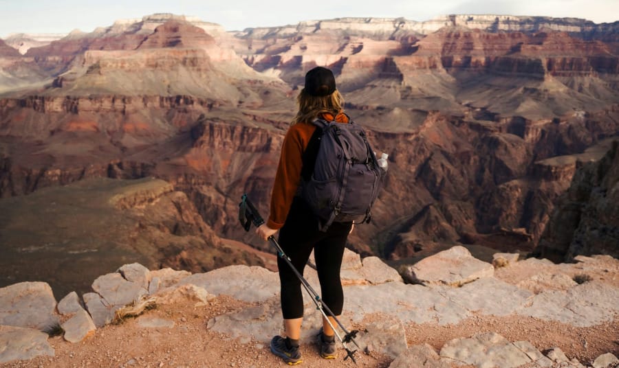 Hiker wearing an orange jacket and backpack stands on a rocky Grand Canyon overlook, holding trekking poles and looking across the canyon.