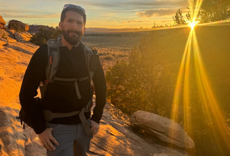 Hiker wearing a black zip-up hoodie and backpack on a sandstone trail at sunset, with golden sunlight illuminating the desert landscape.
