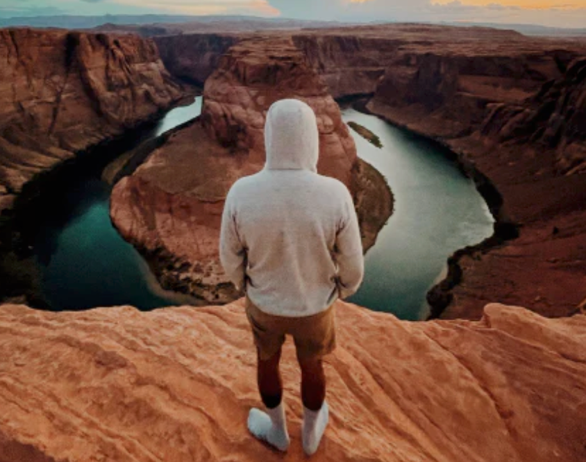 Hiker wearing a light-colored hoodie stands on a sandstone overlook above Horseshoe Bend and the Colorado River in Arizona at sunset.