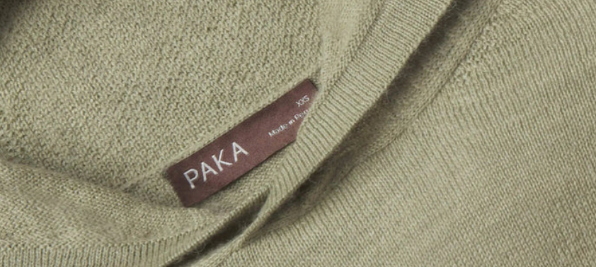 Close-up of sage green PAKA alpaca wool knit fabric showing the sweater's texture, neckline, and PAKA clothing label.
