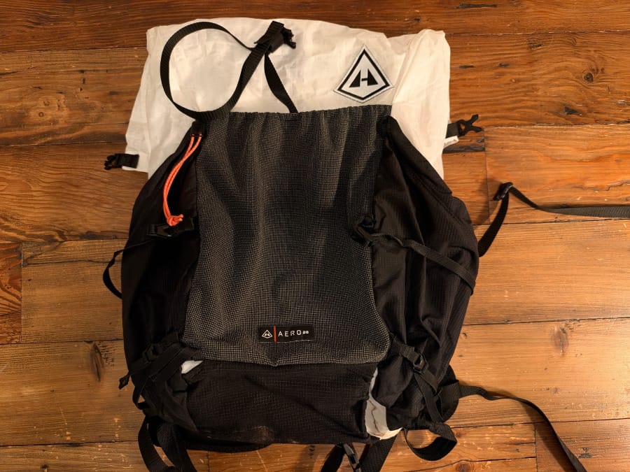 Hyperlite Mountain Gear Aero 28 backpack laid flat on a wooden floor showing its large black mesh exterior pocket, white Dyneema body, side pockets, and compression straps.