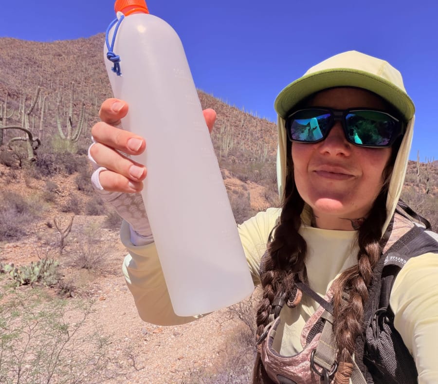 Hiker wearing the Hyperlite Aero 28 backpack while holding a lightweight water bottle on a sunny Sonoran Desert trail surrounded by saguaros.