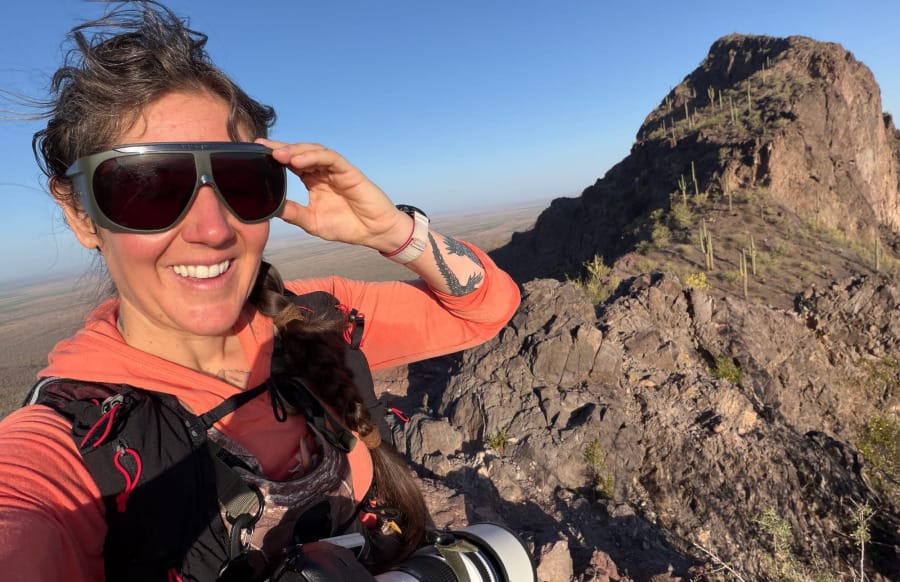 Outdoor photographer wearing a coral-colored hiking hoodie, sunglasses, and a black hiking pack while carrying a telephoto camera on a rocky, saguaro-covered mountain trail in Arizona.