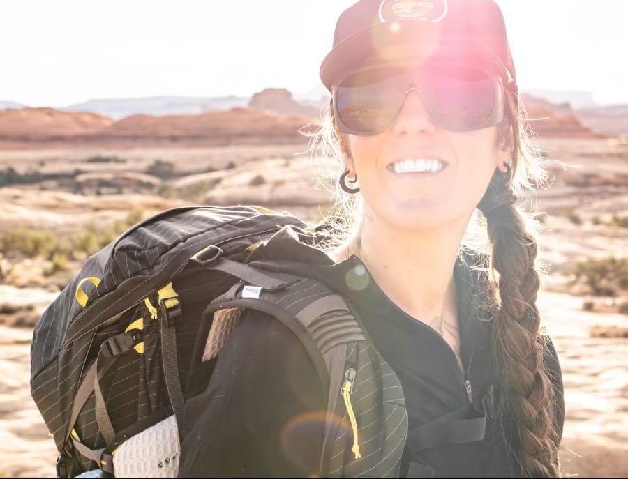 Smiling backpacker wearing a black hiking hoodie, sunglasses, and a cap while carrying a large backpack through a sunlit red rock desert landscape.