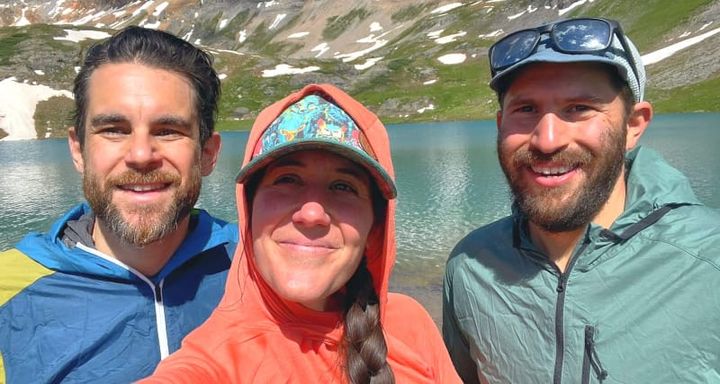  Three hikers wearing sun hoodies smiling in front of a turquoise alpine lake surrounded by rugged mountains and snow patches.