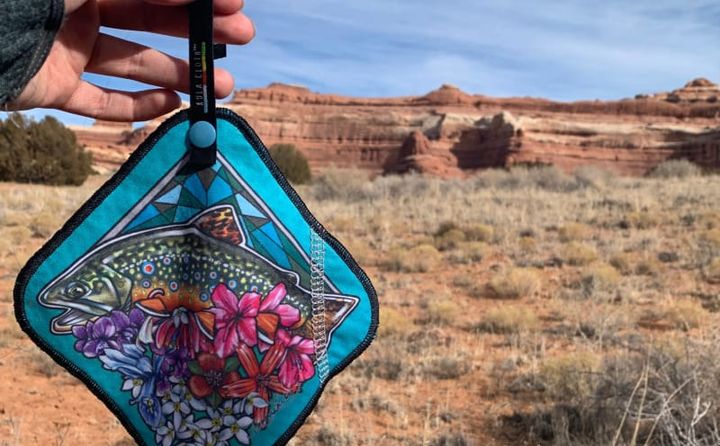  Hand holding a colorful Kula Cloth with a trout and flower design in a dry desert environment with sandstone formations in the background.