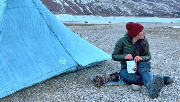 Woman sitting by a Dyneema ultralight tent at a high elevation lake, eating a backpacking meal at sunset.