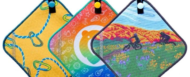 Three Kula Cloths hanging side by side, featuring a rope design, a rainbow design, and a mountain biking desert scene design.