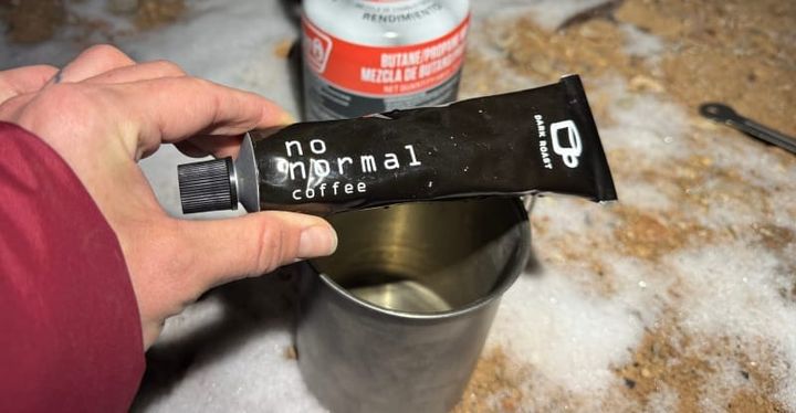  Hand holding No Normal Coffee paste in front of a camp stove and mug in a snowy outdoor setting, showcasing its use for backcountry coffee.
