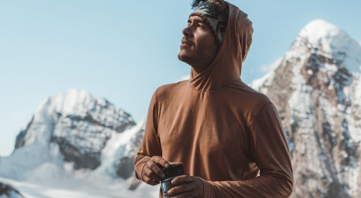  Man wearing a brown PAKA SOL Hoodie and holding a water bottle labeled “Mountains Please,” standing in front of snow-covered peaks during a mountain expedition.
