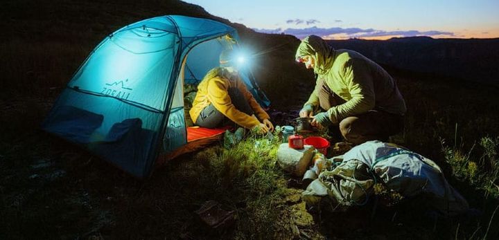 Campers cooking dinner at dusk beside the glowing Zorali Summit-Seeker 2P Tent in the backcountry