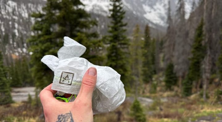 Hand holding Adotec Gear Rain Mitt Stuff Sacks in front of snow-dusted mountains and evergreen trees