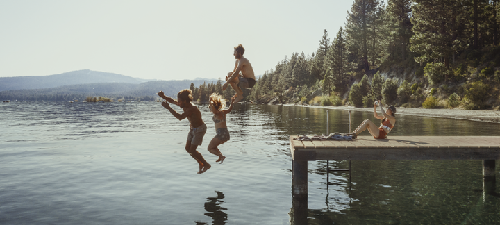  Three people jumping into a mountain lake from a dock while another lounges in a swimsuit, capturing summer joy and outdoor fun—REI 2025 Fourth of July Sale.