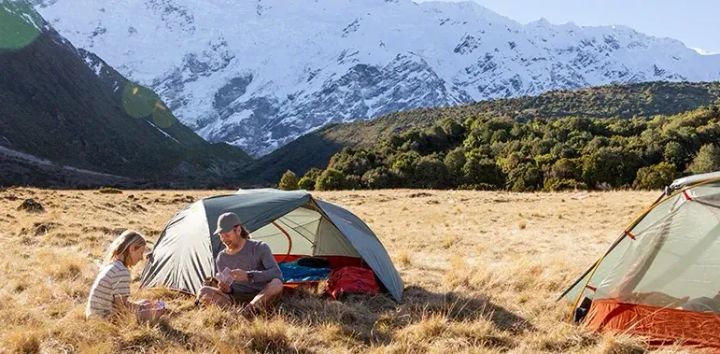  Two people sitting outside a Sea to Summit tent in an alpine meadow with snow-covered mountains in the background, showcasing lightweight backpacking gear in a scenic wilderness setting.